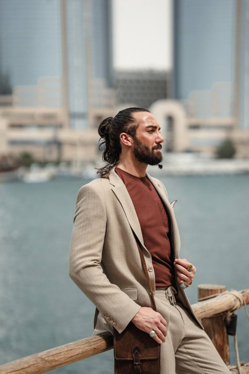 Side view of a man in a light beige Mazonni suit paired with a brown t-shirt and crossbody bag, showcasing relaxed men’s fashion with city skyline backdrop