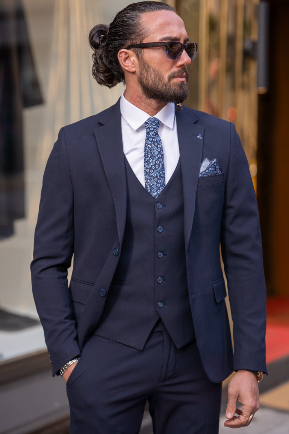 Slim-fit three-piece suit in dark blue by Mazonni, styled with sunglasses and sleek dress shoes for a modern formal look